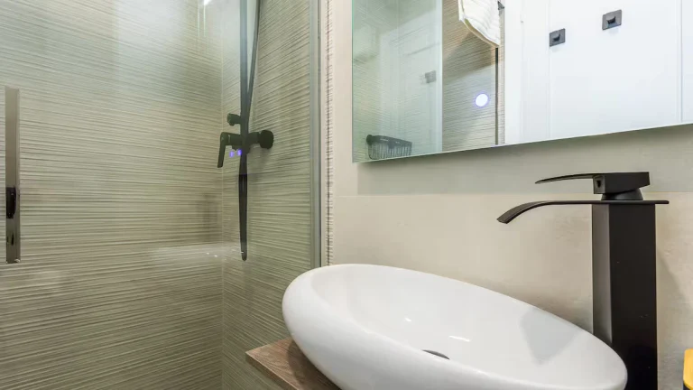 Baño moderno con lavabo ovalado blanco, grifo negro, ducha con vidrio y azulejos beige texturizados.