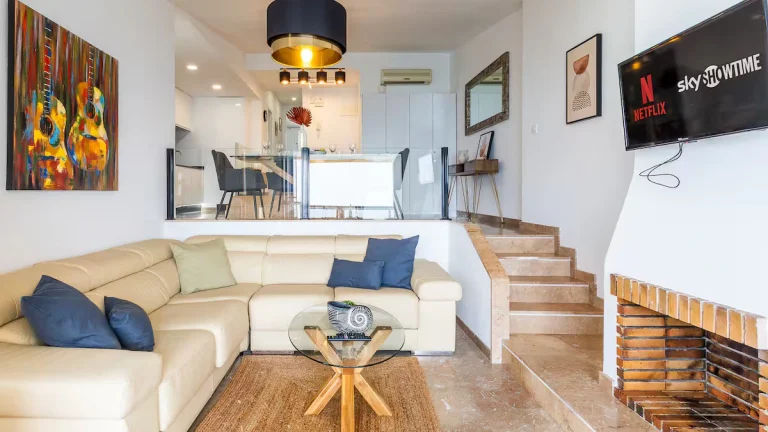 Sala moderna con sofá beige, cojines azules, mesa de vidrio, chimenea, TV y arte de guitarras. Escaleras hacia comedor blanco arriba.