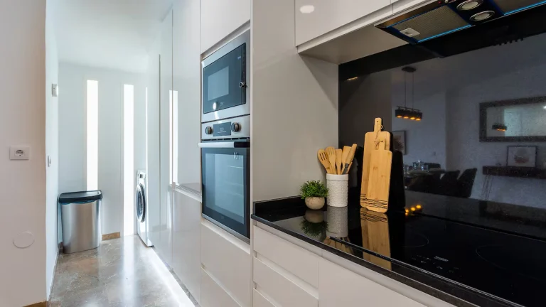 Cocina moderna con electrodomésticos empotrados, estufa negra, utensilios de madera, y una lavadora en el fondo iluminado.