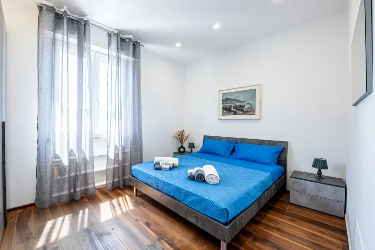 Dormitorio con cama doble cubierta por ropa de cama azul, cortinas grises, lámparas de noche negras y suelo de madera.