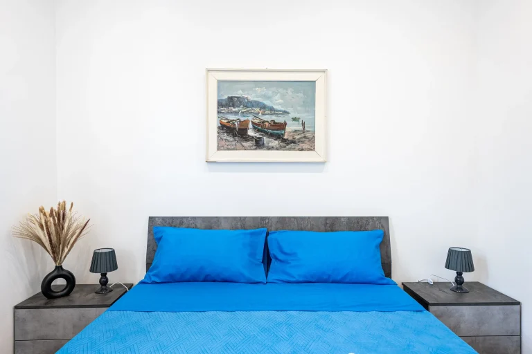 Cama con sábanas azules, lámparas grises en mesas de noche. Cuadro de paisaje costero con barcos sobre la pared blanca.