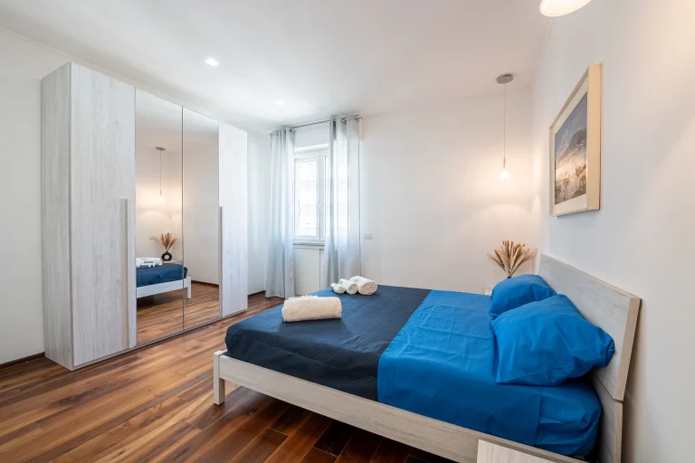 Dormitorio moderno con cama de sábanas azules, armario con puertas espejadas, suelos de madera y luz natural.