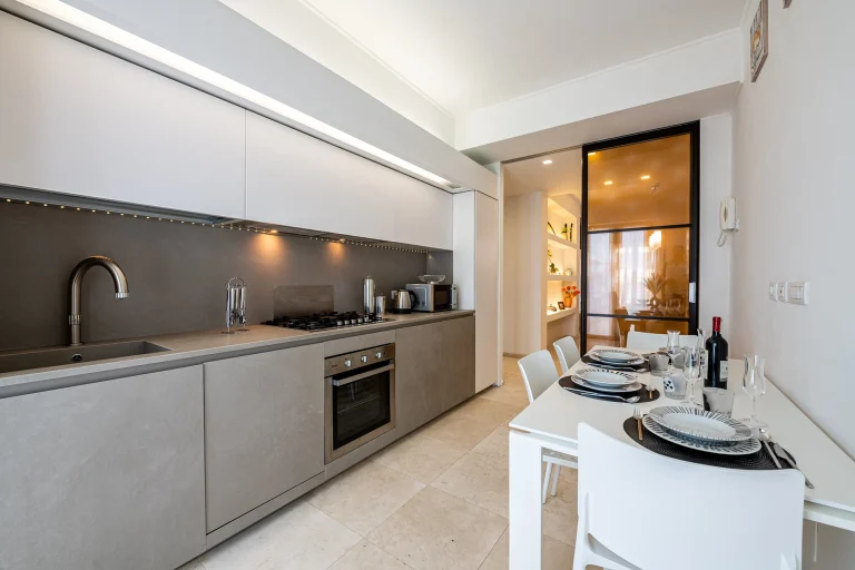 Cocina moderna con encimeras grises, horno empotrado, mesa blanca con platos y copas, luz tenue, estanterías al fondo.