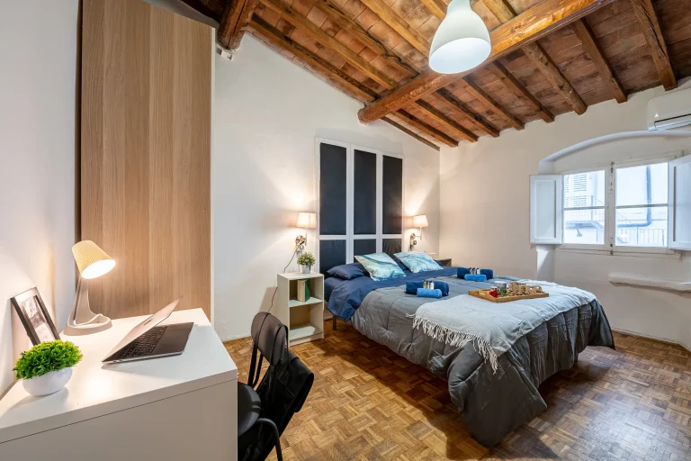 Habitación con techo de vigas de madera, cama doble con mantas gris y azul, escritorio con laptop y silla, ventana amplia.