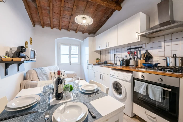 Cocina rústica con techos de madera, mesa puesta para cuatro, electrodomésticos modernos y estantería con cafetera.