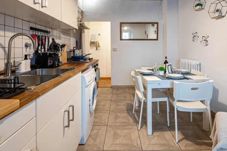 Cocina moderna con encimera de madera, tostadora, lavadora, y mesa blanca con cuatro sillas. Decoración simple y plantas.