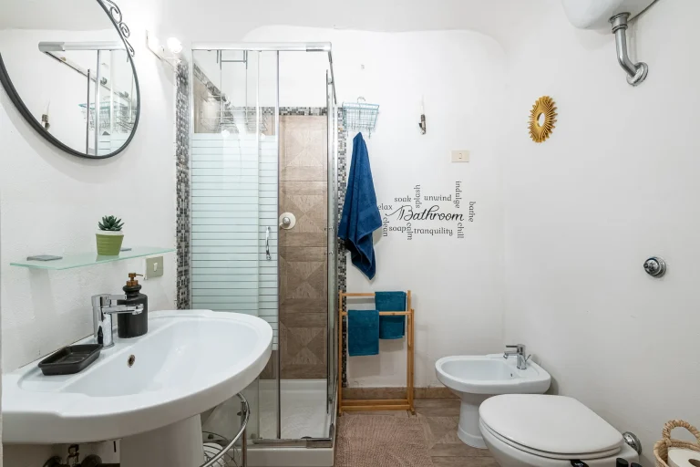 Baño con ducha de vidrio, lavabo blanco, espejo redondo, toallas azules, bidé, decoración de pared y espejo pequeño.