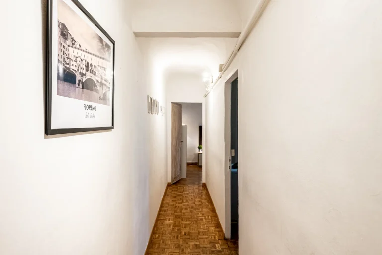 Pasillo estrecho con suelo de parqué, paredes blancas y una imagen enmarcada de Florencia. Luz suave al fondo.