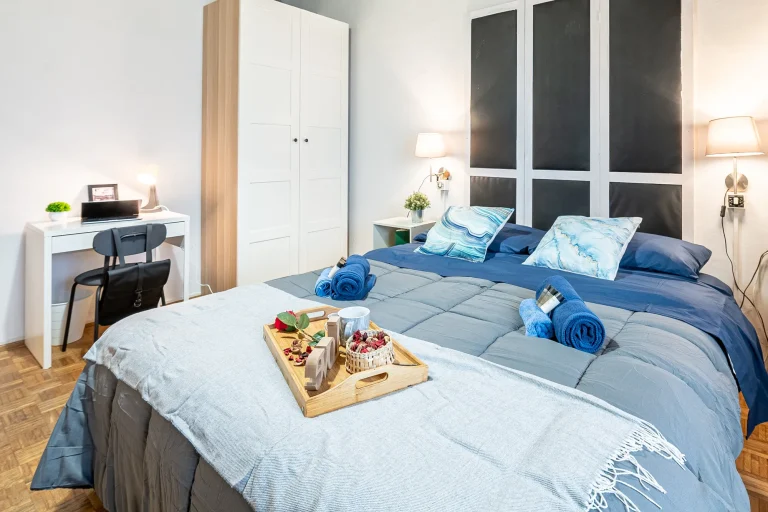 Habitación moderna con cama doble, ropa de cama gris y azul, almohadas decorativas, escritorio blanco, lámparas de pared y una bandeja con desayuno.