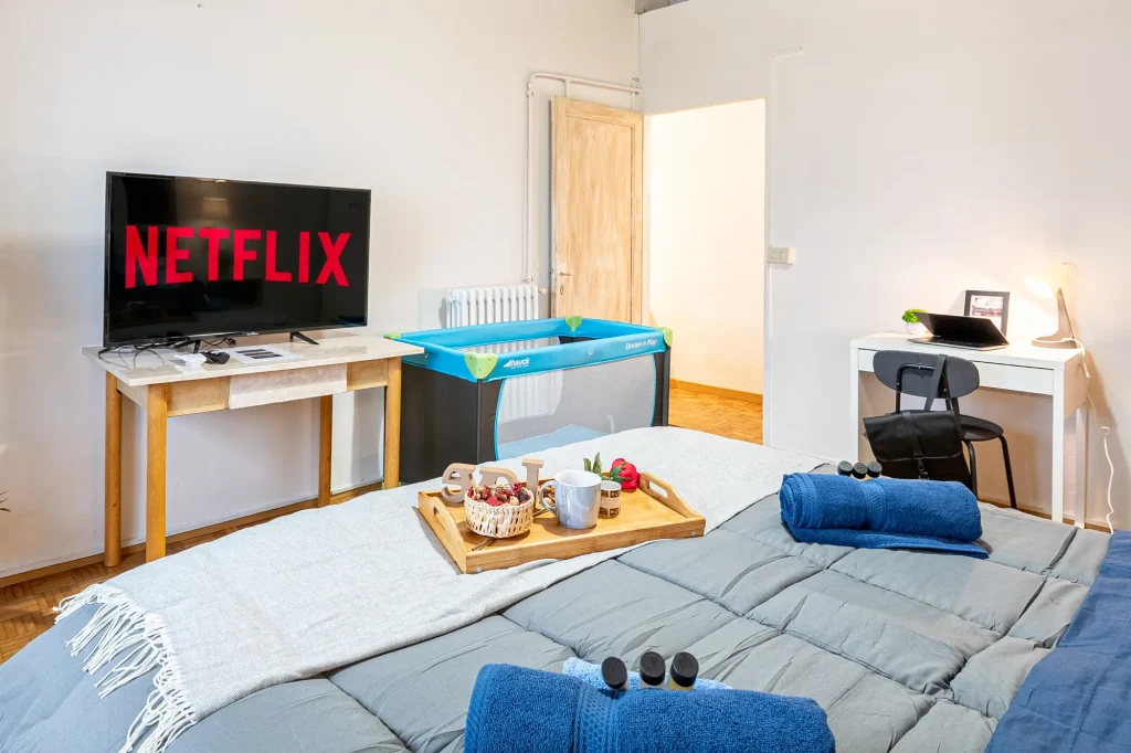 Habitación moderna con cama gris, bandeja con desayuno, televisor con Netflix, cuna portátil, escritorio con lámpara y tablet.