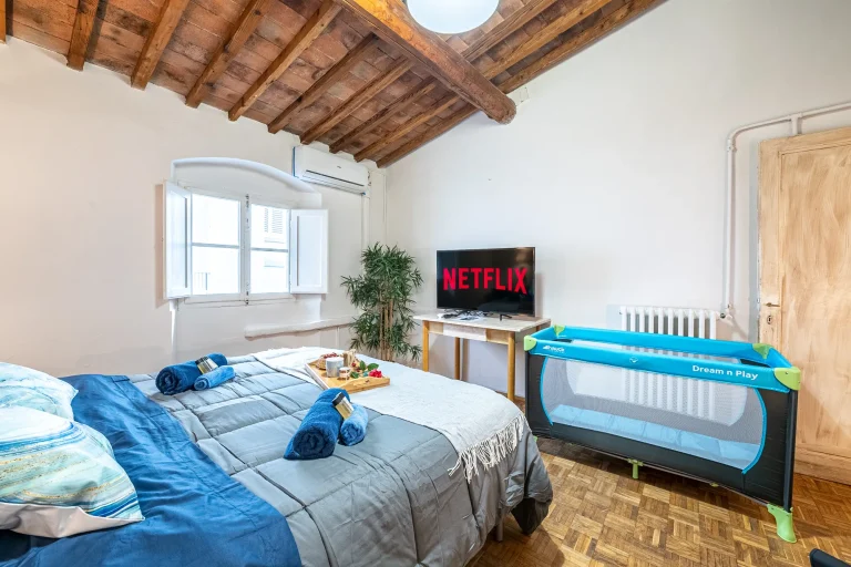 Habitación con cama, edredones azules, TV con Netflix, cuna, planta decorativa, ventana abierta y techo de madera.