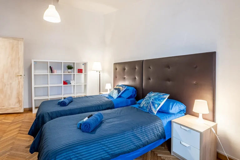 Habitación con dos camas individuales, ropa de cama azul, cabecero acolchado oscuro, lámparas, estantería blanca y suelo de madera.