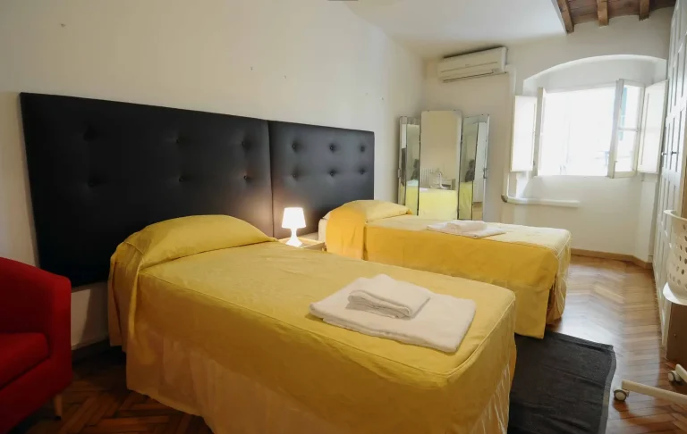 Habitación con dos camas individuales con sábanas amarillas, cabecero negro, lámpara de noche, ventana, espejo y silla blanca.