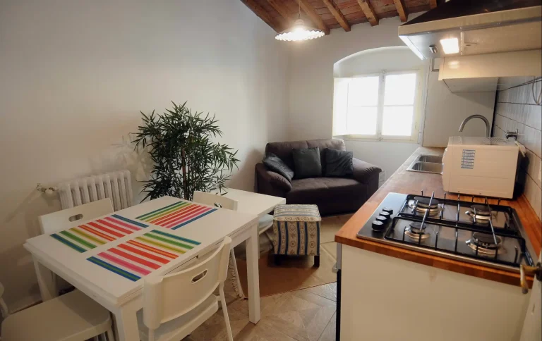 Cocina pequeña con sofá marrón, mesa blanca y sillas. Encimera de madera con estufa de gas y fregadero. Ventana iluminando.