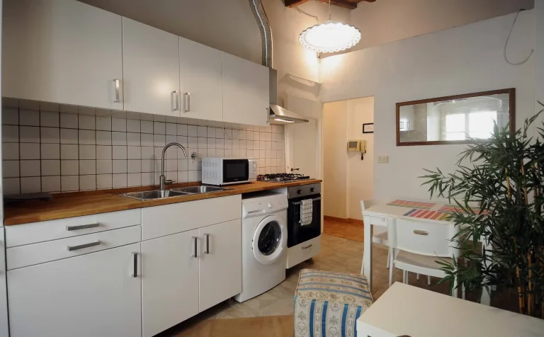 Cocina pequeña con muebles blancos, lavadora, horno, microondas, mesa con mantel colorido y planta en esquina. Lámpara colgante.