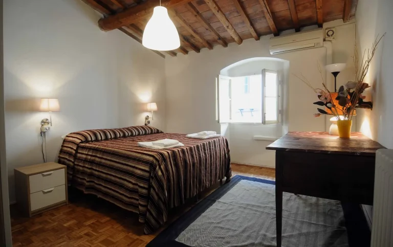 Habitación con cama de rayas marrones, mesitas de noche, ventana abierta, techo de vigas de madera y lámparas de pared.