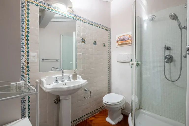 Baño con azulejos decorativos, espejo grande, lavabo clásico, ducha de cristal y toallas. Elementos en blanco y azul.