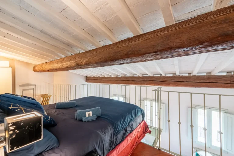 Habitación con techo de vigas de madera, cama con ropa azul, paredes blancas, ventanas con contraventanas y barandilla metálica.