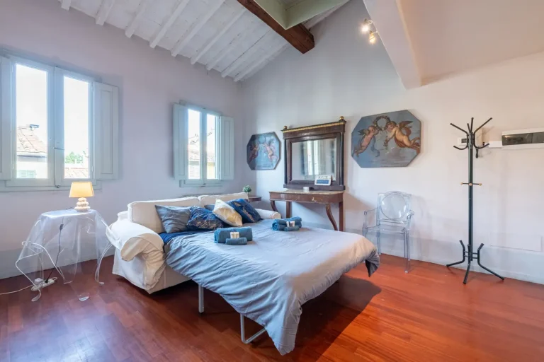 Habitación luminosa con cama, almohadas azules, pinturas de ángeles, espejo sobre cómoda y perchero. Suelo de madera.