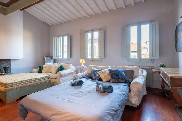 Sala luminosa con cama y sofá blancos, ventanas de madera, techo con vigas, mesa y cojines coloridos.