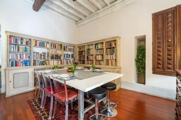 Comedor con mesa blanca, sillas transparentes, librerías con abundantes libros, ventana de madera y techo con vigas expuestas.