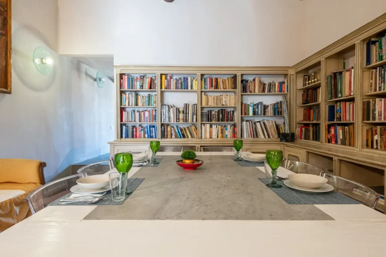 Comedor con mesa de mármol, sillas transparentes, copas verdes, platos blancos y estantería llena de libros al fondo.