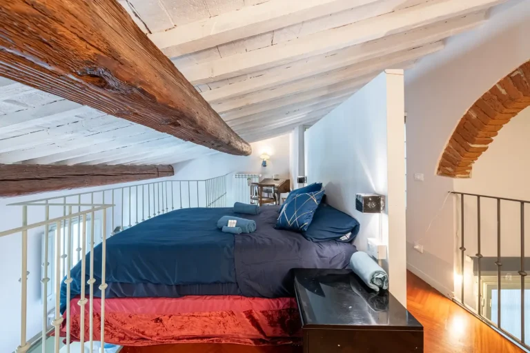 Dormitorio en altillo con cama grande de sábanas azules, vigas de madera expuestas y una pared de ladrillo visto.