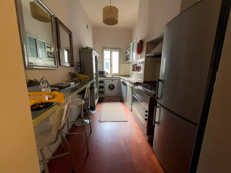 Cocina estrecha con suelo de madera, electrodomésticos de acero, lavadora junto a la ventana y barra con sillas altas.