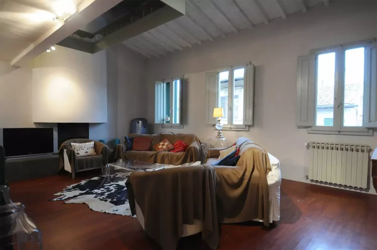 Sala de estar con sofás cubiertos, alfombra de piel de vaca, televisor grande, lámpara encendida y ventanas con vista exterior.