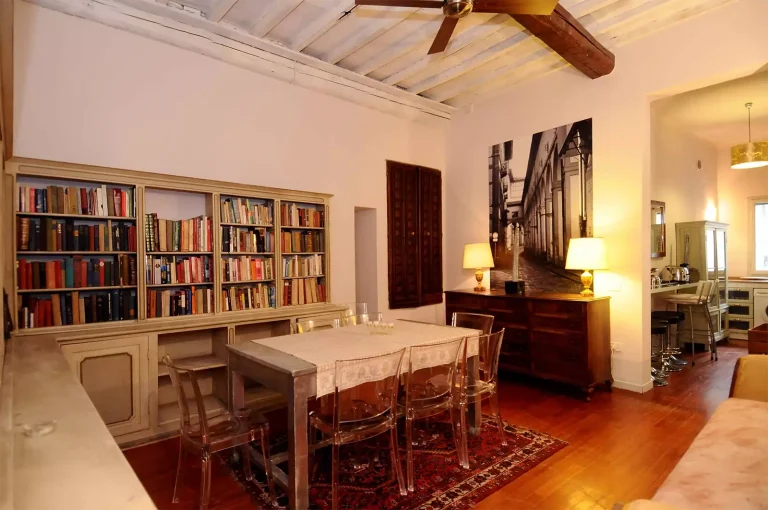 Interior de comedor elegante con estantería llena de libros, mesa de madera, sillas transparentes, lámparas y cuadro en blanco y negro.