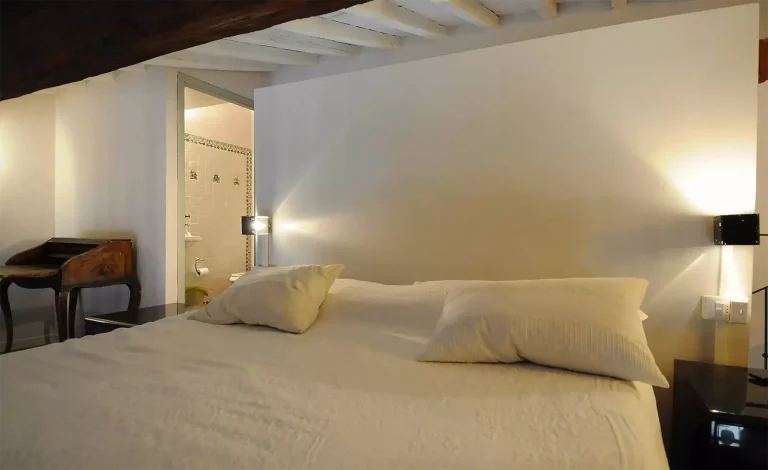 Dormitorio con cama grande, almohadas blancas, mesa de madera, baño visible al fondo, luz cálida y techo de vigas.