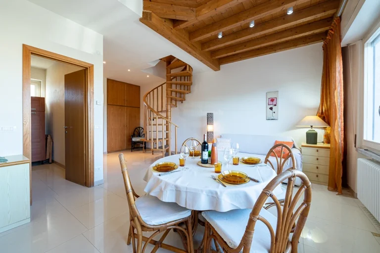 Sala de estar luminosa con techo de madera, escalera de caracol, mesa de comedor con vajilla y sofá con cojines. Ventana amplia con cortinas naranjas.