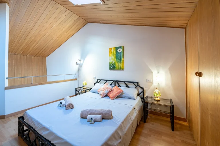 Dormitorio acogedor con techo de madera, cama doble con cabecera negra y sábanas claras. Mesitas de noche con lámparas, armario de madera y barandilla lateral.