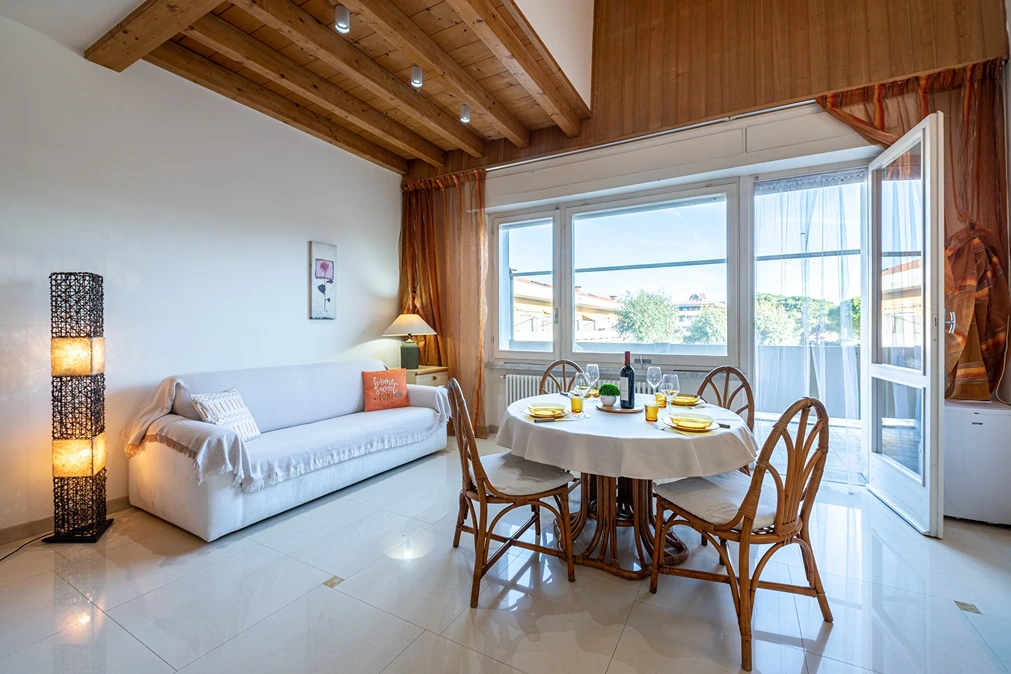 Sala de estar luminosa con techo de madera, sofá blanco y mesa de comedor con vajilla. Ventanales amplios con vista exterior y cortinas naranjas.