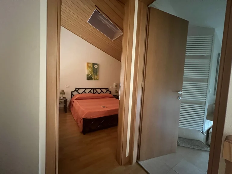 Vista de un pasillo con acceso a un dormitorio y un baño. La habitación tiene cama con colcha naranja, techo de madera y mesitas de noche. El baño cuenta con toallero calefactable.