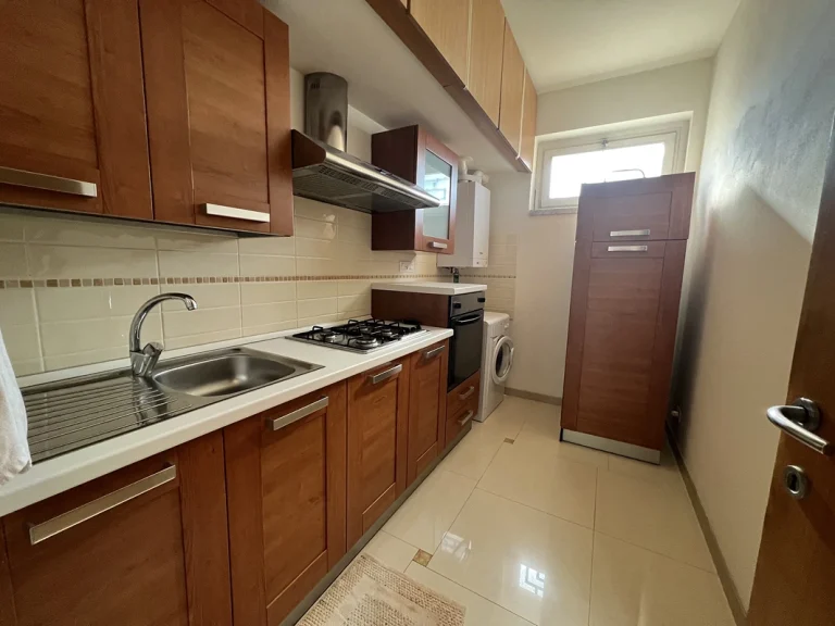 Cocina estrecha con muebles de madera, encimera blanca y azulejos beige. Fregadero, estufa con campana extractora, horno, lavadora y ventana pequeña.