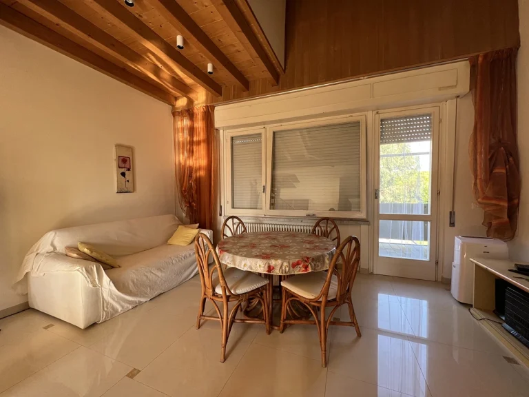 Sala con iluminación cálida, muebles de madera clara y decoración rústica. Mesa con sillas de mimbre, televisor pequeño y puerta con acceso al exterior.