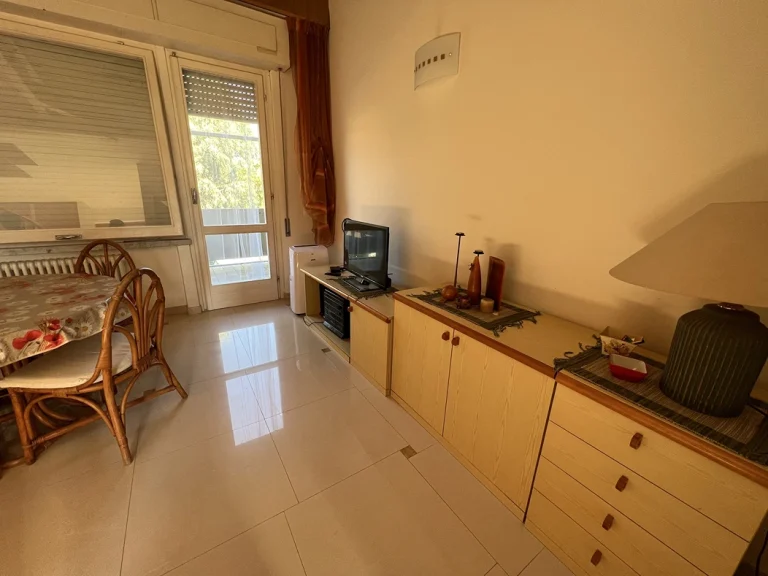 Sala con iluminación cálida, muebles de madera clara y decoración rústica. Mesa con sillas de mimbre, televisor pequeño y puerta con acceso al exterior.