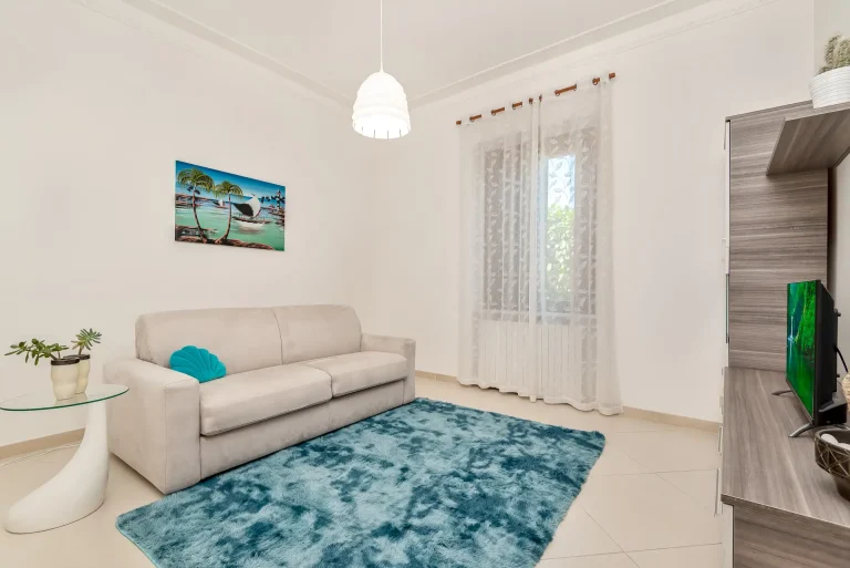 Sala de estar luminosa con sofá beige, alfombra azul y mesa auxiliar de vidrio. Decoración con cuadro tropical, cortinas blancas y estantería con TV.