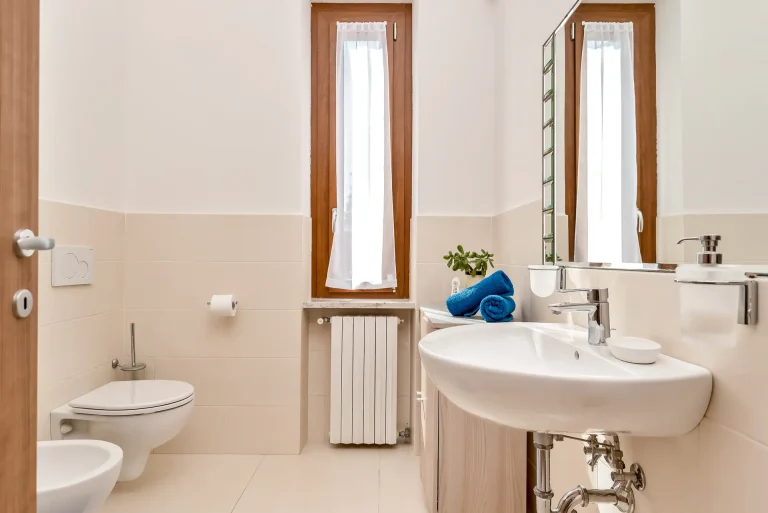 Baño luminoso con inodoro suspendido, lavabo amplio y espejo grande. Ventanas con cortinas blancas, toallas azules dobladas y detalles en madera.