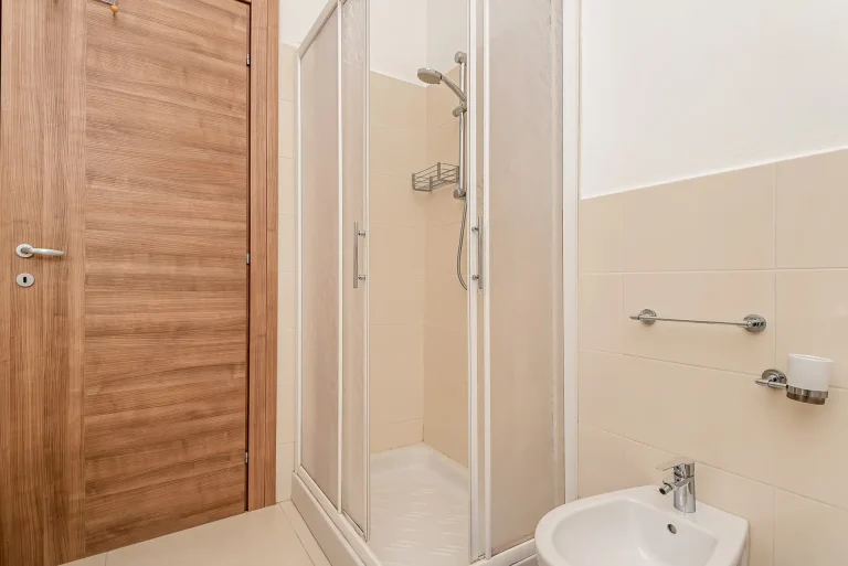 Baño con ducha de mampara corrediza, lavabo blanco y pared revestida con azulejos beige. Puerta de madera clara y accesorios metálicos en la pared.