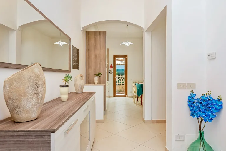 Pasillo luminoso con muebles de madera clara, espejo grande y decoración en cerámica. Al fondo, puerta de vidrio con acceso al exterior y mesa con mantel azul en la cocina.