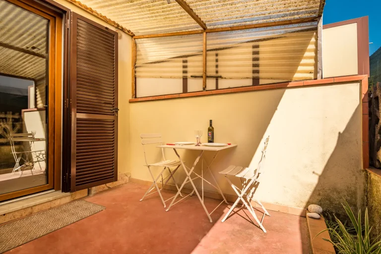 Pequeña terraza con suelo de cemento rojo, mesa blanca con dos sillas y vajilla con botella de vino. Puerta de madera con contraventana y techo de policarbonato.