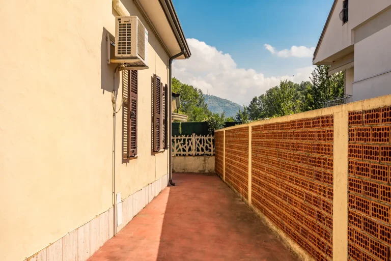 Pasillo exterior con suelo de cemento rojo, pared de ladrillos y vista a colinas arboladas. Fachada beige con ventanas de contraventanas oscuras y aire acondicionado.