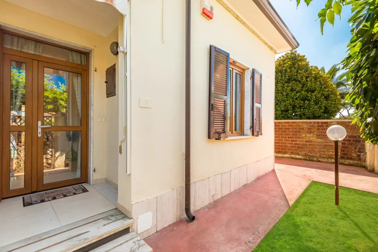Entrada de casa con puerta de madera y vidrio, escalones de mármol y sendero de cemento. Ventana con contraventanas oscuras y jardín con césped y farola.
