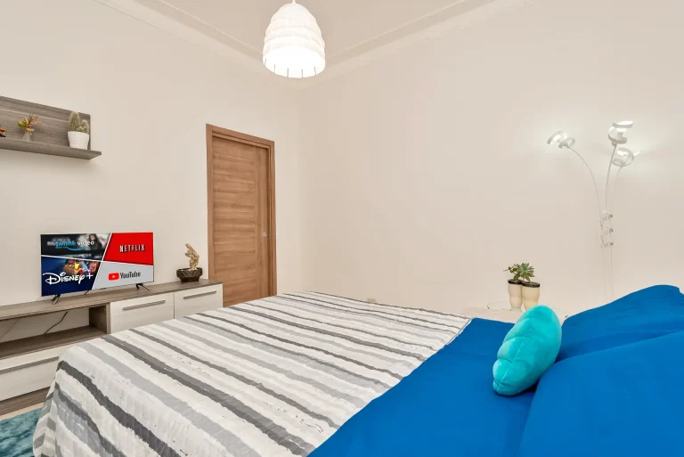 Dormitorio moderno con cama de sábanas azules y colcha a rayas. Cojín turquesa, televisor con streaming, lámpara colgante y decoración minimalista.