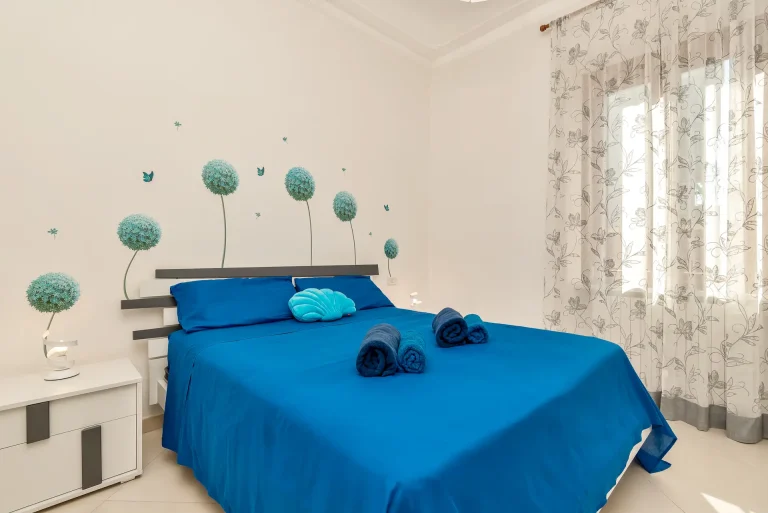 Habitación con cama de sábanas azul intenso, cojín turquesa y toallas enrolladas. Decoración con mural de flores y mariposas. Cortinas translúcidas.