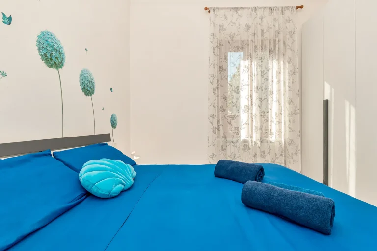 Dormitorio con cama de sábanas azul oscuro, cojín en forma de concha turquesa y toallas enrolladas. Decoración con mural floral y cortina translúcida.