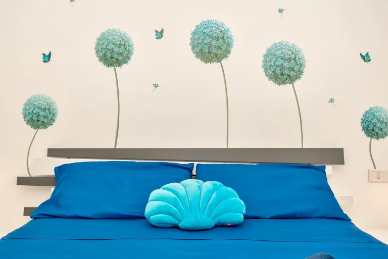 Cama con sábanas y almohadas azul oscuro, cojín en forma de concha turquesa y mural decorativo con flores y mariposas en tonos celestes.