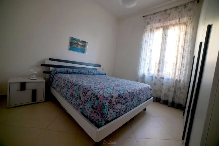 Habitación luminosa con cama de diseño moderno, edredón estampado floral, mesa de noche minimalista y cortinas claras con motivos florales.